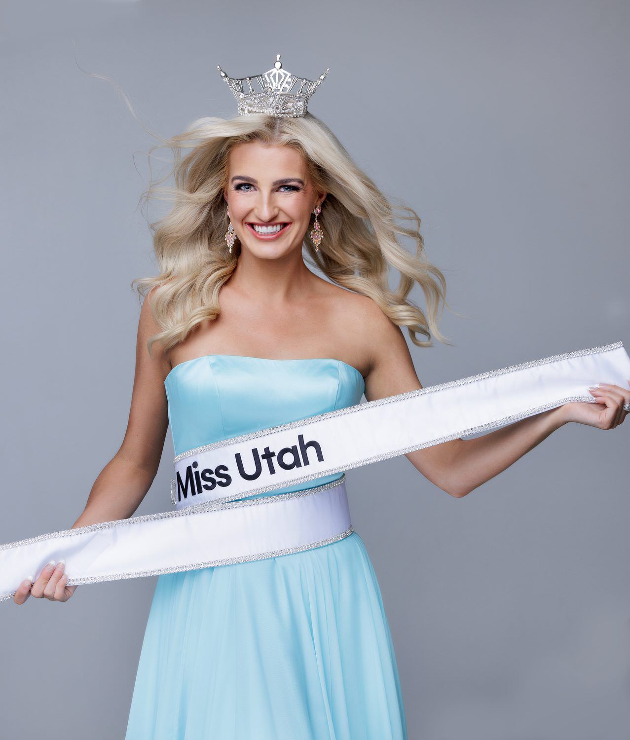 Miss Utah in a pale blue gown holding her sash out wide, hair in motion